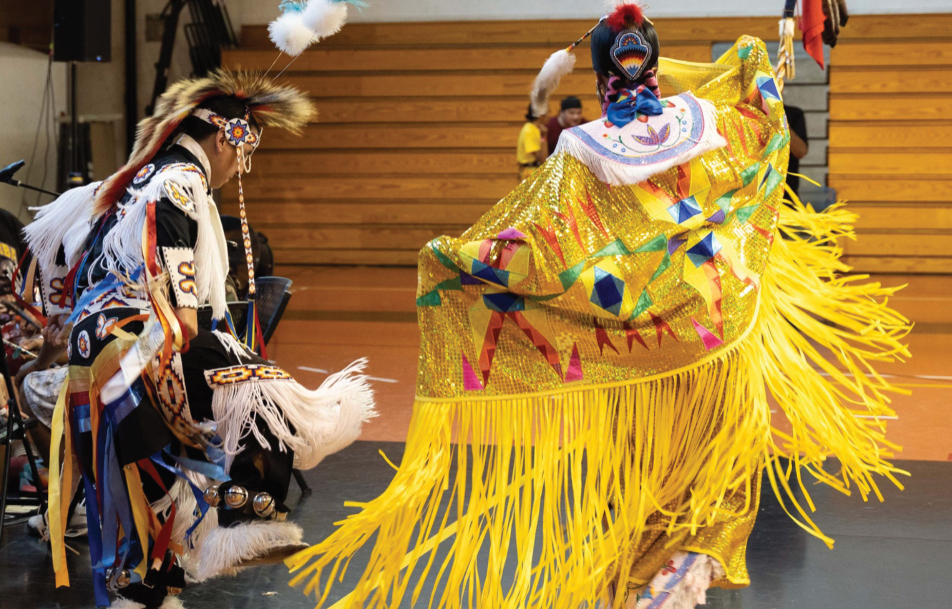 A male and female dressed in regalia dance in a gym.