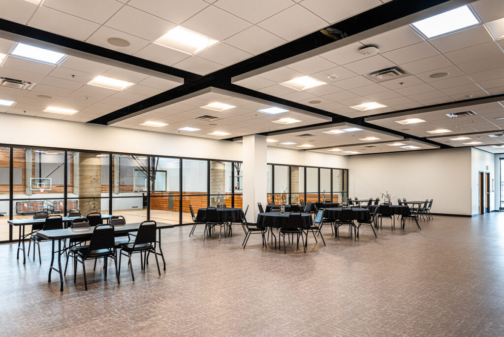 A room with big glass windows overlooking a gym. The room is filled with round tables with chairs.