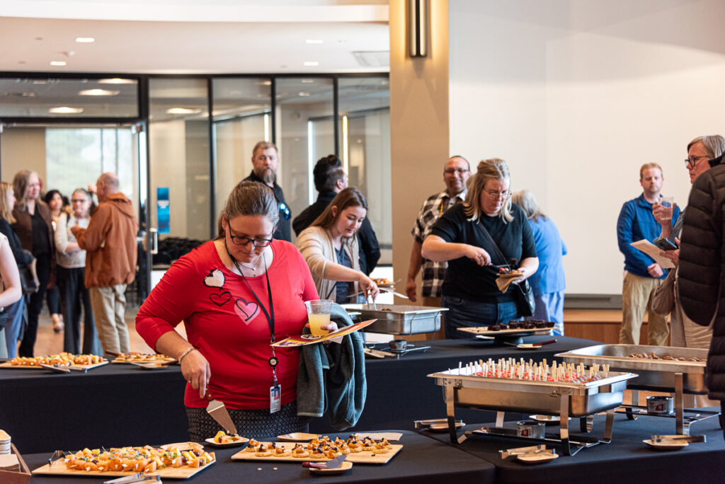 People stand behind a table full of food and use tongs to put food on their plates.