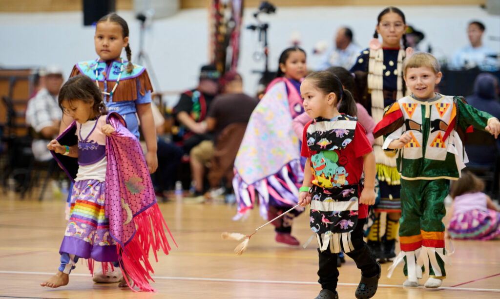 Kids wearing regalia dance in a gym.