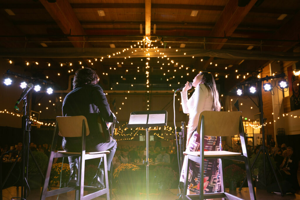 A Native woman and man sing and play guitar in front of an audience in a decorate gym.